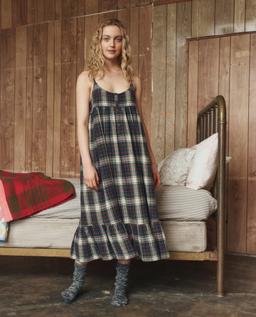 A woman stands beside a bed in a rustic room, wearing THE GREAT. H25 SLEEP’s The Flannel Ruffle Tank Night Dress in Pine Lodge Plaid, paired with patterned socks. The bed features a striped mattress, white pillow, and plaid blanket.