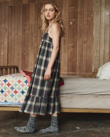 A woman with wavy blonde hair stands indoors in THE GREAT. H25 SLEEP The Flannel Ruffle Tank Night Dress—Pine Lodge Plaid, beside a bed with a colorful quilt and wooden headboard.