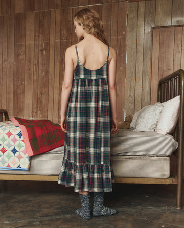 A woman wearing THE GREAT. H25 SLEEP’s Flannel Ruffle Tank Night Dress in Pine Lodge Plaid stands with her back to the camera beside a bed with patterned blankets in a rustic wood-paneled room.