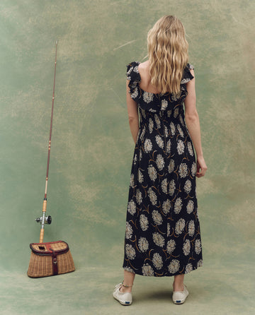 A woman with wavy blond hair wears THE GREAT. SU25's The Havana Dress in Black Dancing Floral and white sneakers, standing near a fishing pole and wicker basket against a mottled green backdrop.