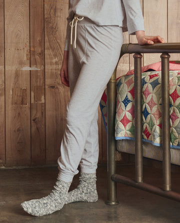 A person stands indoors wearing THE GREAT. H25 SLEEP The Long John in Heather Grey, paired with thick gray socks, beside a metal-framed bed topped with a colorful patchwork quilt.