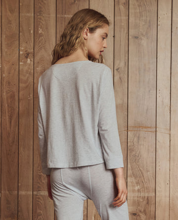 A person with wavy blond hair wears THE GREAT. H25 SLEEP’s The Crop Sleep Henley in Heather Grey and matching pants, standing facing a wooden plank wall with their head turned slightly to the side.