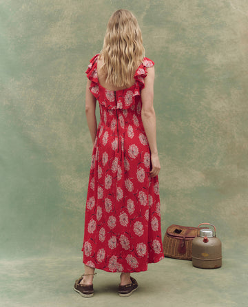 A woman with wavy blonde hair faces away, wearing THE GREAT. SU25's The Havana Dress in True Red Dancing Floral, paired with brown sandals. Vintage containers sit at her feet against a green backdrop.