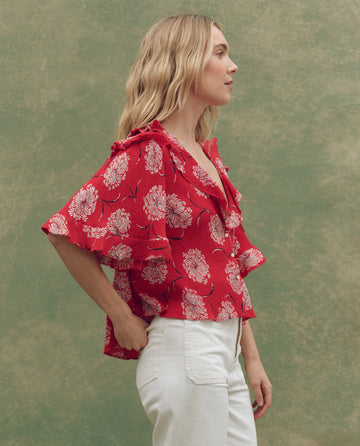 A woman stands in profile wearing THE GREAT. SU25 The Bower Top in True Red Dancing Floral with ruffled sleeves and white pants, set against a mottled green background, highlighting chic women's fashion.