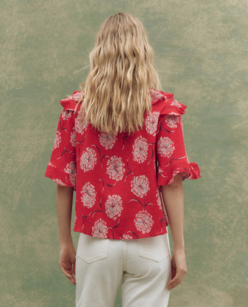 Seen from behind, a woman with long, wavy blonde hair wears THE GREAT. SU25's The Bower Top in True Red Dancing Floral with ruffled sleeves and white pants against a mottled green background—an elegant take on women’s fashion.
