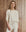 A person with long, wavy blonde hair wears THE GREAT. H25 SLEEP's The Crop Sleep Henley in Washed White, paired with matching cotton drawstring pants, standing before a wooden plank wall.