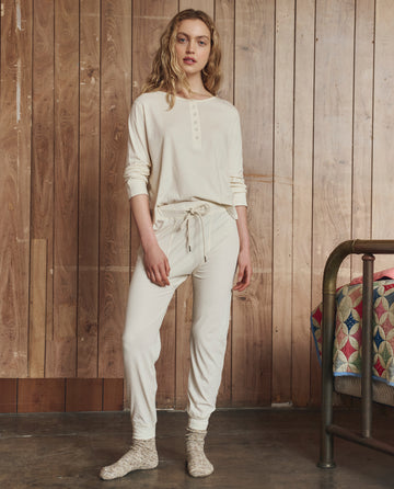 A person stands indoors wearing THE GREAT. H25 SLEEP’s The Crop Sleep Henley in Washed White with matching 100% cotton joggers and socks, beside a bed topped with a colorful patchwork quilt.