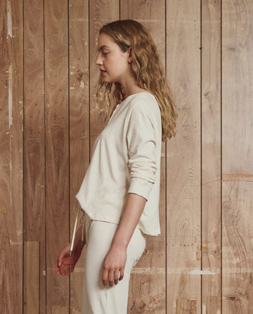 A woman with long, wavy blonde hair stands in profile against a wooden plank wall, wearing THE GREAT. H25 SLEEP The Crop Sleep Henley in Washed White and matching pants, both crafted from 100% cotton.