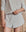 A person wearing THE GREAT. H25 SLEEP Tap Short in Heather Grey with a gathered waistband and a light grey long-sleeve top, standing against a wooden background—ideal for cozy nights.