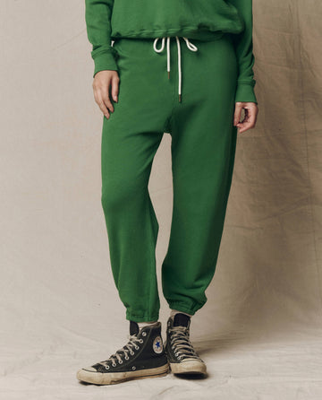 Someone wears The Stadium Sweatpant in Solid Fennel by THE GREAT., styled with a matching green sweatshirt and black Converse sneakers, standing on a neutral backdrop for a fresh take on athletic wear.