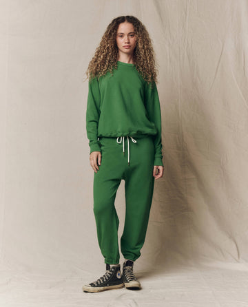 A person with long curly hair wears THE GREAT. SU26 SEASONAL CORE’s The Stadium Sweatpant in Solid Fennel and a matching green sweatshirt with white drawstrings, paired with black high-top sneakers, against a beige fabric backdrop.