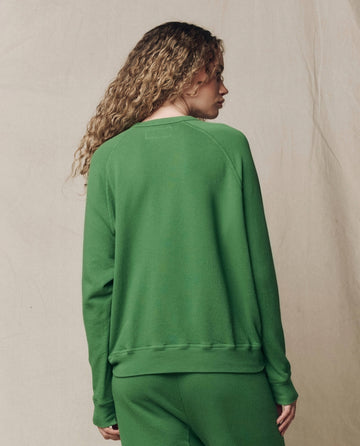 Person with curly hair wears THE GREAT.'s The College Sweatshirt in Solid Fennel, SU26 SEASONAL CORE, and matching pants, standing with their back to the camera against a neutral backdrop.