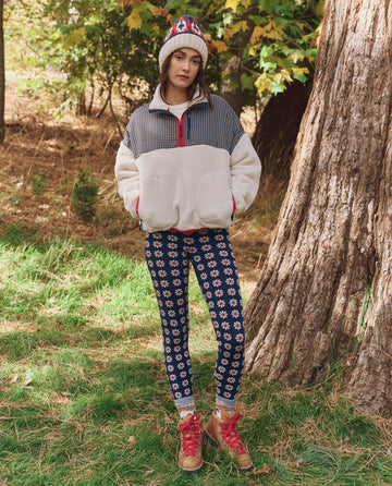 A woman stands outdoors by a tree wearing THE GREAT. F25 TGO The Print-Mix Plush Terrain Half-Zip in Mini Frosted Daisy & Cream, paired with floral leggings, wool socks, a patterned beanie, and hiking boots.
