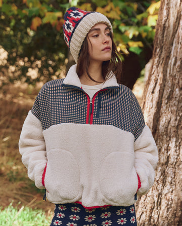 Wearing THE GREAT. F25 TGO's Print-Mix Plush Terrain Half-Zip in Mini Frosted Daisy & Cream, a person stands outdoors in front of a tree and foliage, pairing the cozy patterned jacket with a knit hat and patterned pants.