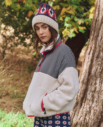A woman stands outdoors near a tree, wearing THE GREAT. F25 TGO The Print-Mix Plush Terrain Half-Zip in Mini Frosted Daisy & Cream, with greenery and fallen leaves in the background.