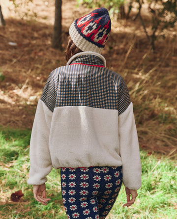 Wearing THE GREAT. F25 TGO Print-Mix Plush Terrain Half-Zip in Mini Frosted Daisy & Cream, someone strolls on grass among trees, showcasing the jacket’s cozy style and playful pattern.