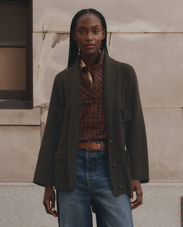 A person stands against a stone wall wearing THE GREAT. F26 D1 The Commander Riding Cardigan in Army, featuring leather knot buttons, paired with a plaid shirt, blue jeans, and a brown belt; their hair is braided and their expression is neutral.