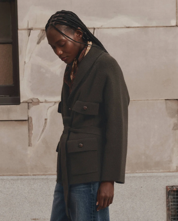 Standing outdoors against a stone wall, the person wears THE GREAT. F26 D1′s The Commander Riding Cardigan in Army—a dark belted jacket featuring large pockets and distinctive leather knot buttons—paired with blue jeans, looking down with braided hair.