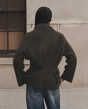 A person stands with their back to the camera, wearing THE GREAT. F26 D1 The Commander Riding Cardigan in Army and blue jeans, in front of a beige wall with a window on the left.