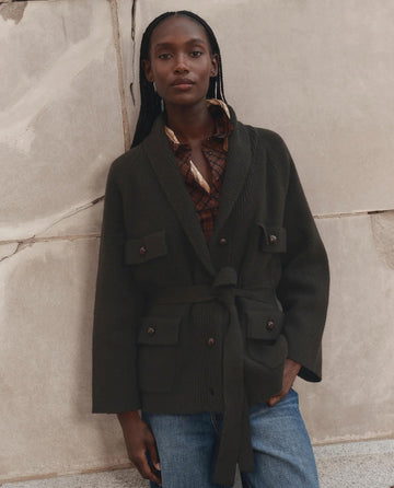 A person stands against a stone wall, hands in pockets and long hair styled in braids, wearing THE GREAT. F26 D1′s The Commander Riding Cardigan in Army with distinctive leather knot buttons, layered over a plaid shirt and blue jeans.