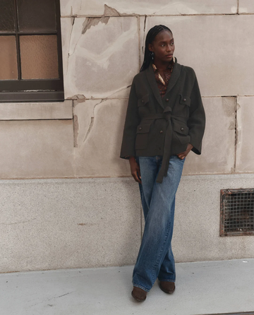 A person stands against a stone wall wearing THE GREAT. F26 D1 The Commander Riding Cardigan in Army, styled with wide-leg jeans, brown shoes, and a patterned shirt, with one hand in their pocket.