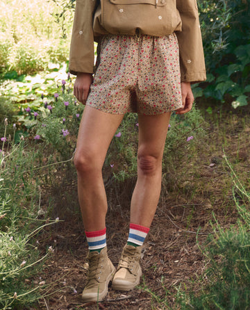 A person outdoors, surrounded by greenery and wildflowers, wears THE GREAT. SP26 TGO’s The Rover Short in Sprouting Floral, paired with a tan jacket, striped socks, and tan hiking boots.