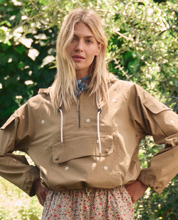 A woman with long blonde hair stands outdoors in THE GREAT. SP26 TGO’s Embroidered Cropped Trailhead Pullover—khaki with daisy embroidery—and a floral skirt, with green foliage in the background.