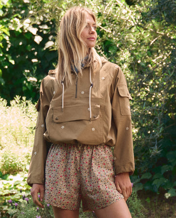 Amid lush greenery, a person wears THE GREAT. SP26 TGO's The Rover Short in Sprouting Floral, paired with a tan hooded jacket featuring floral embroidery.
