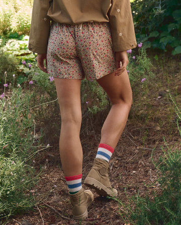 A person stands outdoors in greenery, seen from behind, wearing THE GREAT. SP26 TGO's The Rover Short in Sprouting Floral with a tan long-sleeve shirt, striped socks, and tan boots.