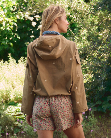 A person stands outdoors with their back to the camera, surrounded by greenery, wearing THE GREAT. SP26 TGO’s Embroidered Cropped Trailhead Pullover in khaki with daisy embroidery and floral shorts.