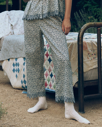 A person wearing The Great. SP26 Sleep Eyelet Easy Sleep Pant in Bluebell Ditsy stands by a metal-frame bed with a colorful quilt outdoors, paired with white socks.