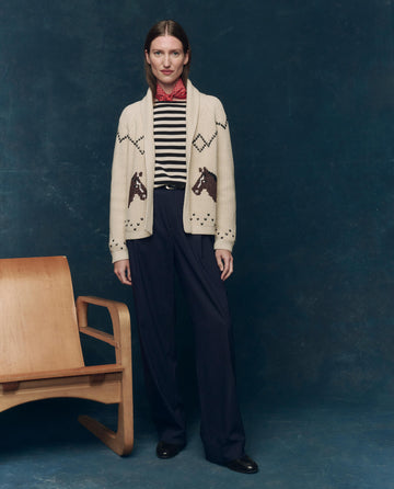 A person stands indoors next to a wooden chair, wearing THE GREAT. SP25 COLLECTORS The Horse Lodge Cardigan in Cream, a striped shirt, pinstripe trousers, and black shoes against a dark blue backdrop.