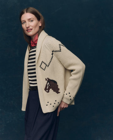 A person stands sideways against a dark backdrop, wearing THE GREAT. SP25 COLLECTORS The Horse Lodge Cardigan in cream, featuring horse and zigzag designs, over a black-and-white striped shirt, navy pants, and a red neck scarf.