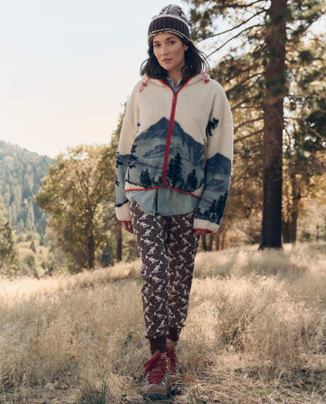 A woman outdoors wears THE GREAT. F26 TGO The Plush Horizon Full Zip. -- Snowcap View Landscape, with patterned pants, red boots, and a knit hat amid grassy hills and trees.