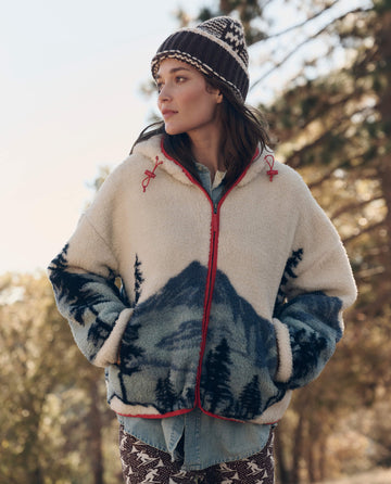 A woman outdoors in The Plush Horizon Full Zip. -- Snowcap View Landscape by THE GREAT. F26 TGO, with patterned pants and a knit hat; sunlight shines through trees behind her.