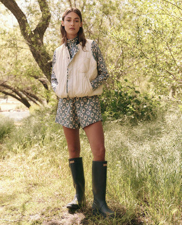 Surrounded by lush green grass and trees, a person stands outdoors wearing a patterned outfit, rubber boots, and The Reversible Ultra Light Down Vest in Falling Daisy and Cream from THE GREAT. SP25 TGO.
