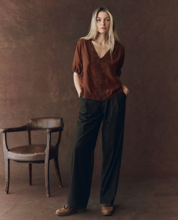 A woman stands indoors beside a wooden chair, wearing THE GREAT. F25 D1 The Short Sleeve Legacy Top in Chocolate, black wide-leg pants, and brown shoes against a brown textured backdrop.