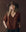 A woman with long blonde hair wears THE GREAT. F25 D1’s The Short Sleeve Legacy Top in Chocolate and a wide-brimmed brown hat, standing against a dark brown background with her hands in her pockets.