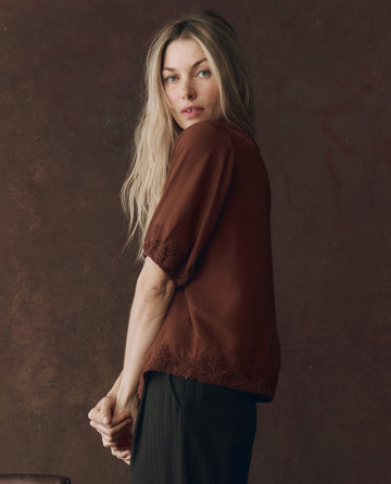 A woman with long blonde hair stands against a dark brown background, wearing THE GREAT. F25 D1’s The Short Sleeve Legacy Top in Chocolate and dark pants, looking toward the camera.