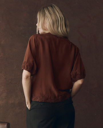 A person with blonde hair, seen from behind, wears THE GREAT. F25 D1 The Short Sleeve Legacy Top in Chocolate and dark pants against a textured brown background.