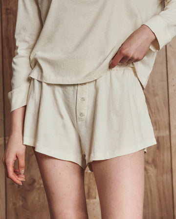 A person wearing THE GREAT. H25 SLEEP’s The Tap Short in Washed White, a loose 100% cotton sleep short, paired with a matching long-sleeve top, stands against a wooden background.