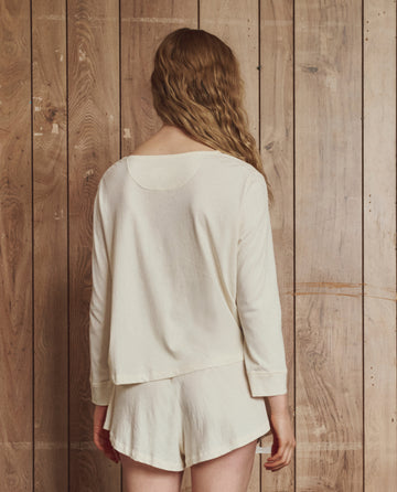 A person with long wavy hair, wearing a loose cream top and THE GREAT. H25 SLEEP The Tap Short.. in Washed White, made from 100% super soft cotton jersey, stands facing a wooden wall.