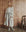 A woman stands indoors on a concrete floor, wrapped in THE GREAT. H25 SLEEP The Sweatshirt Robe in Light Heather Grey, with gray socks and a light shirt, as a patchwork quilt rests on a brass bed beside her.