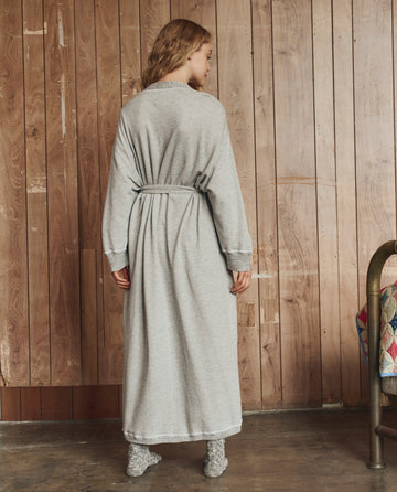 A person stands indoors facing a wooden wall, wearing THE GREAT. H25 SLEEP The Sweatshirt Robe in Light Heather Grey and gray socks, hands by their sides, head turned slightly to the right.