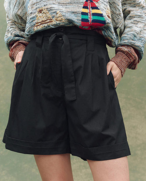 Person wearing THE GREAT. SU25 The Pleated Trouser Short in black, with a tie belt, hands in pockets, styled with a boat-patterned sweater, standing against a green background.