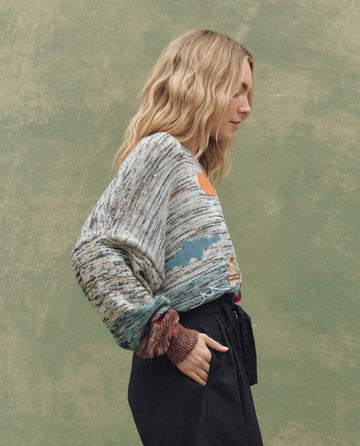 A woman with long blonde hair models THE GREAT. SU25 The Boating Pullover in Sea Breeze Multi and black high-waisted pants, standing in profile before a muted green background.