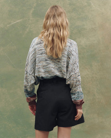 A person with long, wavy blonde hair faces away, wearing THE GREAT. SU25 The Boating Pullover in Sea Breeze Multi and black shorts against a green textured background.