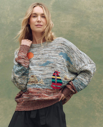 A woman wears THE GREAT. SU25 The Boating Pullover – Sea Breeze Multi, featuring a seaside scene with a sailboat and sun, standing before a mottled green background.