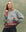 A woman wears THE GREAT. SU25 The Boating Pullover – Sea Breeze Multi, featuring a seaside scene with a sailboat and sun, standing before a mottled green background.