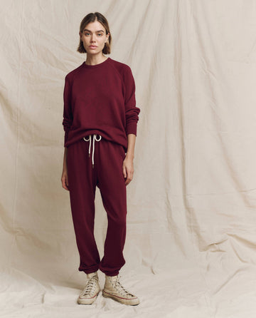 A person wearing The College Sweatshirt in Solid Plum by THE GREAT. HOL 24 KNITS and matching sweatpants, paired with white sneakers, stands against a beige fabric backdrop.